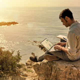 A young man uses his laptop and notepad while working remotely with the sea in the background as the sun sets