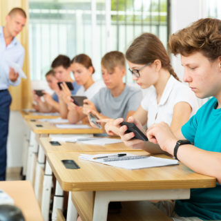 Focused teenager with group of fellow students sitting at lesson in classroom, using mobile phone